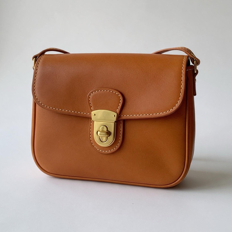 A small tan leather crossbody bag with a brass clasp and cream stitching, alone against a plain light background.